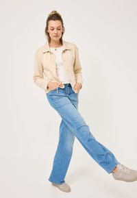 Giacca crema abbottonata sopra una camicia bianca, abbinata a jeans azzurri chiari e sneaker beige. Tessuto morbido con una vestibilità rilassata e gamba dritta.
