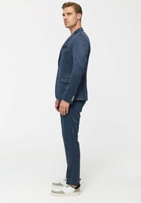 Man in blue suit and white sneakers standing in profile against plain white background.