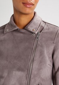 Giacca da moto in suede grigia chiara con colletto stand, bottoni a pressione argentati e chiusura con zip diagonale. Texture liscia con dettagli minimi.