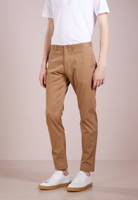 Beige cotton chinos with a slim fit, featuring side pockets and a clean design, paired with white sneakers on a neutral background.