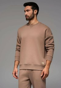 Beige bomullströja med ribbade mudd och nederkant, rund halsringning och en liten logotyp på den nedre sidan. Mjuk textur med en avslappnad passform.