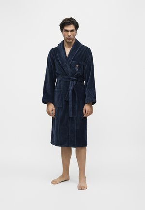 Homme debout pieds nus portant un peignoir en peluche bleu marine avec une ceinture et un ours brodé sur la poche poitrine sur un fond uni.