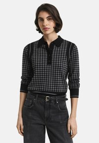 Vrouw in een zwart-grijze houndstooth bedrukte longsleeve poloshirt, ingestopt in hoge zwarte jeans met een zwarte riem.