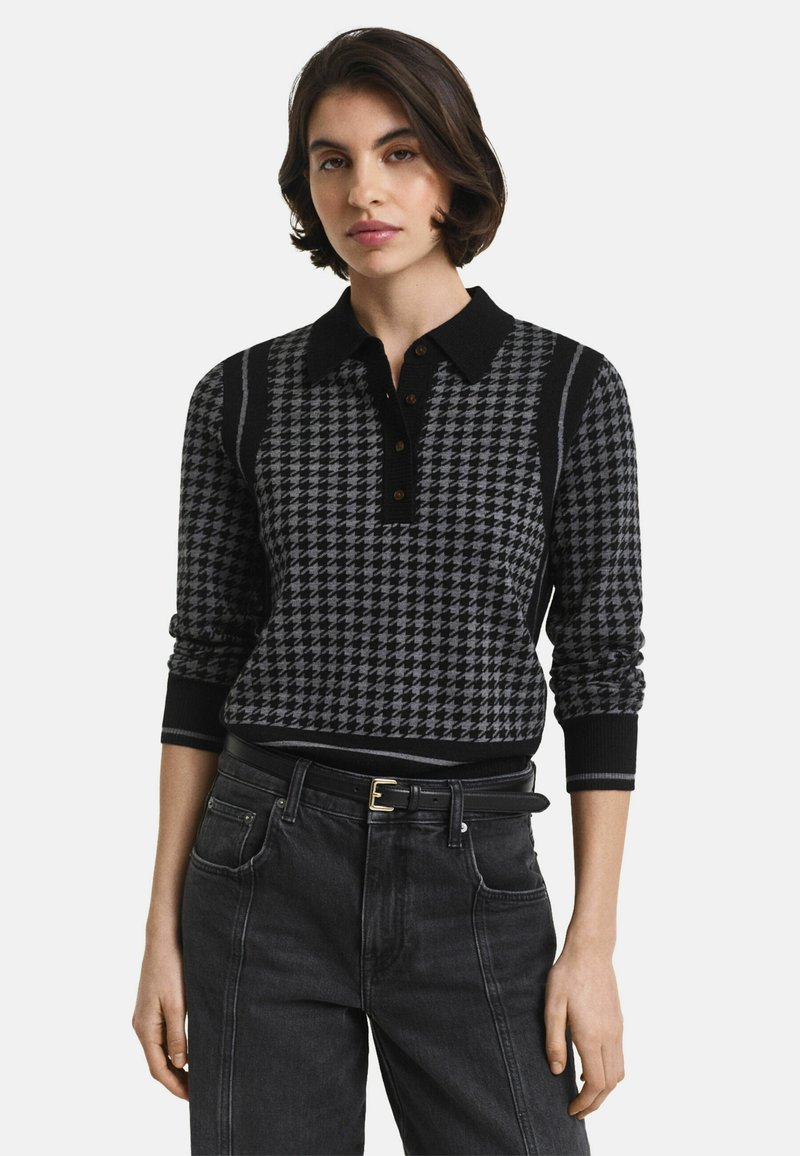 Vrouw in een zwart-grijze houndstooth bedrukte longsleeve poloshirt, ingestopt in hoge zwarte jeans met een zwarte riem.