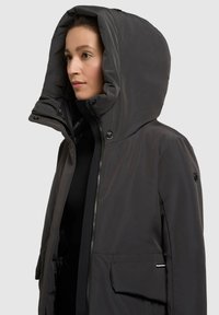 Giacca nera con cappuccio, dalla texture liscia, con zip frontale e bottoni a pressione. Presenta un ampio cappuccio e tasche frontali. Design moderno e slanciato.