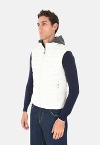 Uomo con un giubbotto bianco trapuntato senza maniche sopra una camicia a maniche lunghe blu navy e jeans blu, che guarda a sinistra su uno sfondo neutro.