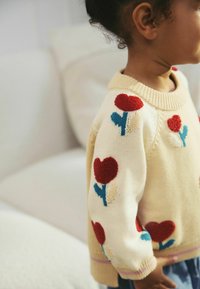 Dzianinowy sweter w kremowym kolorze, z czerwonym sercem i niebieskimi wzorami łodyg.  Żebrowany dekolt i mankiety z różowymi akcentami w szwie.
