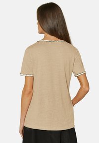 Bruine katoenen t-shirt met korte mouwen, voorzien van een witte en zwarte franjes langs de halslijn en de randen van de mouwen.
