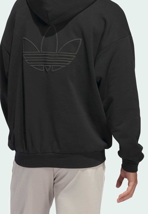 Persoon die een zwarte hoodie draagt met een reliëf Adidas trefoil-logo op de rug en lichtbeige broek, met de rug naar de camera gekeerd.
