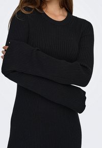 Femme portant une robe en maille côtelée noire avec de longues manches larges, croisant les bras devant un fond blanc uni.