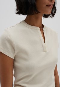 Prążkowany, kremowy top z krótkim rękawem, z rozciętym kołnierzem i luźnym krojem; gładka tekstura podkreśla prostotę w designie.