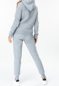 Personne portant un sweat à capuche gris clair et un pantalon de survêtement assorti, debout de dos, baskets blanches visibles.