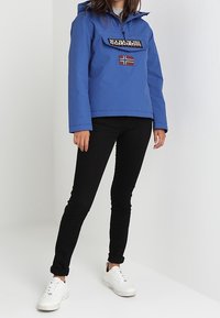 Osoba nosící modrou kapucňovou bundu Napapijri s norskou vlajkou, černé úzké kalhoty a bílé tenisky, stojící na bílém pozadí.