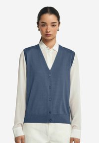 Mörkblå ärmlös cardigan med V-hals, knäppning framtill och ribbade kanter, lager på lager över en vit skjorta med knäppte ärmslut.