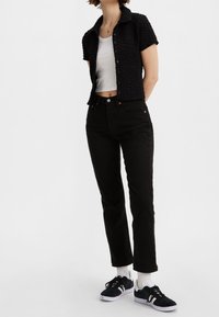 Personne portant une chemise noire texturée à manches courtes boutonnée, un crop top blanc, un jean noir, des chaussettes blanches et des baskets noires avec des rayures blanches.
