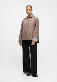 Striped brown shirt with button closure, featuring embroidered white floral accents on wide cuffs, worn with black wide-leg pants.