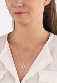 Silverhalsband med hjärtformad berlock som har en mindre hjärtutsnitt, med en strukturerad design, på en delikat kedja framför en ljus blus.