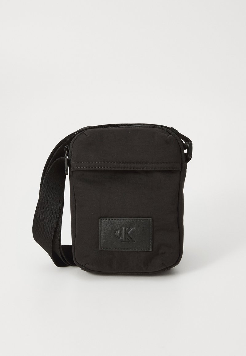 Črna tkaninska torbica čez ramena z zadrgo, sprednjim žepom in usnjenim detajlom s logotipom "cK". Vključena nastavljiva naramnica.