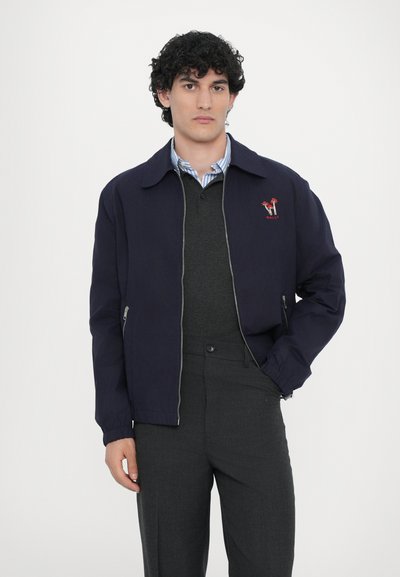 Giacca leggera navy con chiusura zip, design con colletto, polsini elasticizzati e dettaglio logo ricamato rosso, indossata sopra un maglione grigio.