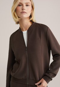 Ženska s kratkimi blond lasmi, oblečena v rjavo jakno z zadrgo čez belo majico, stoji s roko na bokih pred nevtralnim ozadjem.