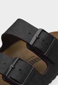 Κοντινό πλάνο μαύρου σανδαλιού με δύο ρυθμιζόμενους ιμάντες και τετράγωνες μεταλλικές αγκράφες πάνω σε καφέ πάτο.