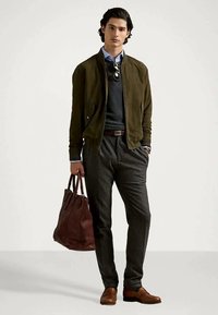 Man wearing a green bomber jacket, blue shirt, dark sweater, gray trousers, brown shoes, and holding a large brown leather tote bag.
