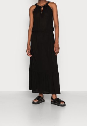 Femme portant une robe maxi noire sans manches avec un col noué, associée à des sandales à plateforme noires, sur un fond blanc uni.