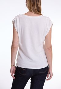 Wit top met korte mouwen en geplooide schouderdetails, zachte textuur, ronde halslijn en een relaxed fit, gecombineerd met donkere spijkerbroek.