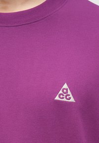 Μωβ φούτερ με στρογγυλή λαιμόκοψη και μικρό λευκό τρίγωνο λογότυπο με τα γράμματα "a", "c" και "g" στην αριστερή πλευρά του στήθους.