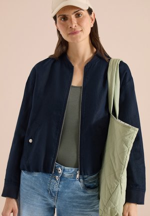 Woman wearing beige cap, navy zip-up jacket, olive green top, light blue jeans, carrying light green quilted tote bag on shoulder.