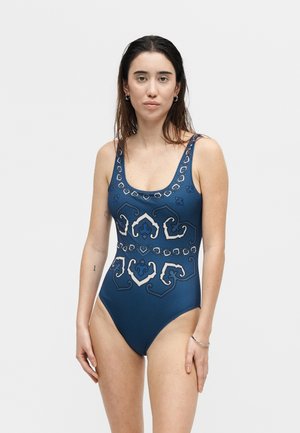 Young woman with long dark hair wears a blue one-piece swimsuit with intricate white and dark blue geometric patterns, standing against a plain background.