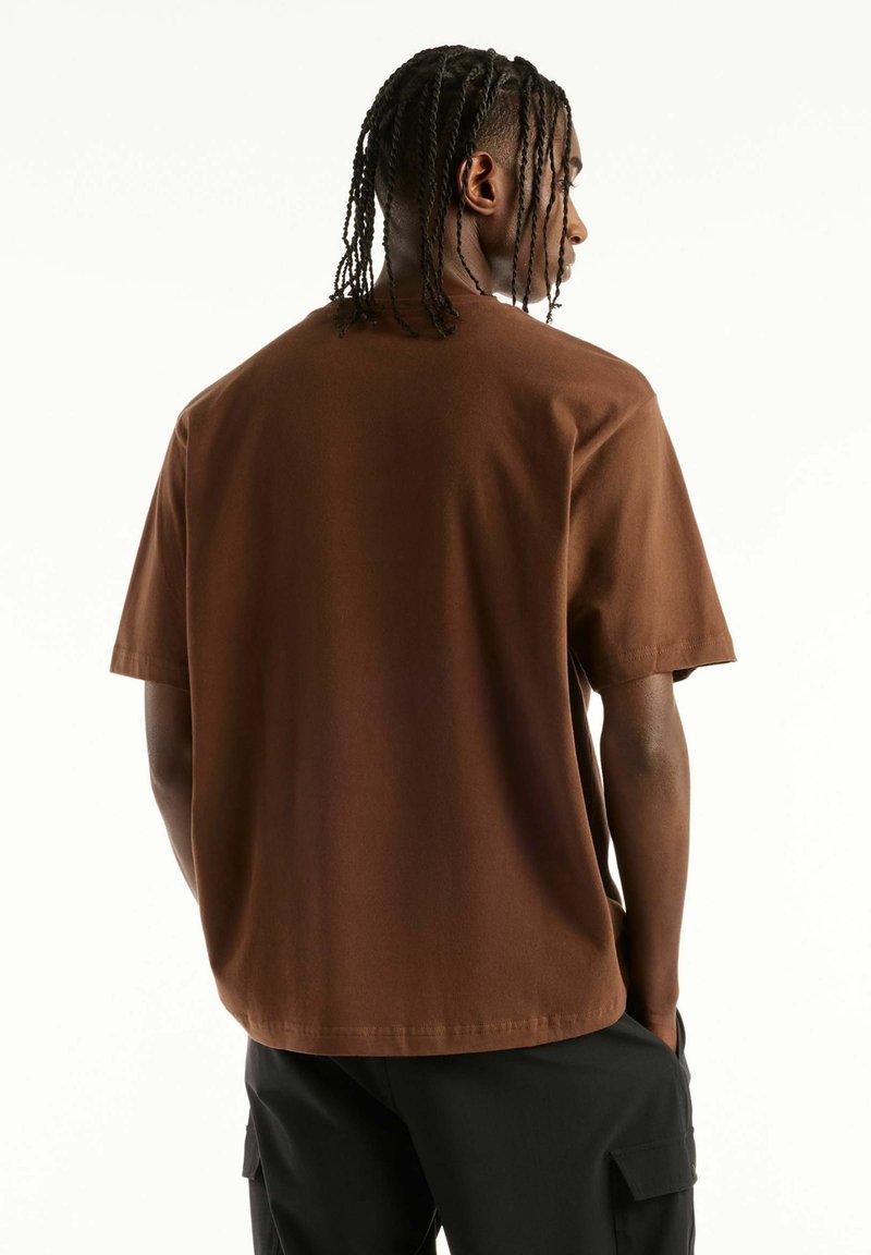 Man with braided hair facing away, wearing a loose brown short-sleeve shirt and black cargo pants against a plain white background.