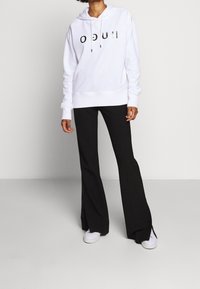 Sudadera blanca con el estampado negro "HUGO", combinada con pantalones negros de campana y zapatillas blancas. Tejido suave con corte relajado y capucha ajustable.