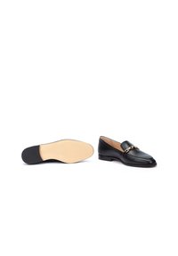 Mocasines de cuero negro con suela plana, que presentan una cadena dorada decorativa en la parte frontal. Textura suave y forma clásica.