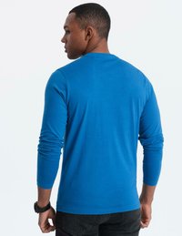 Man som vänder ryggen mot kameran, iklädd en klarblå långärmad skjorta och svarta jeans, med en svart klocka på vänster handled mot en enfärgad bakgrund.