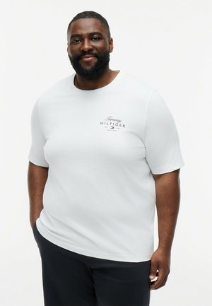 Lächelnder Mann mit Bart, der ein weißes Tommy Hilfiger T-Shirt und schwarze Hose trägt, steht mit einer Hand in der Tasche vor weißem Hintergrund.