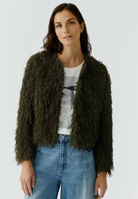 Olive green, shaggy-textured jacket with a cropped design, featuring a round neckline and open front, worn over a graphic t-shirt and jeans.