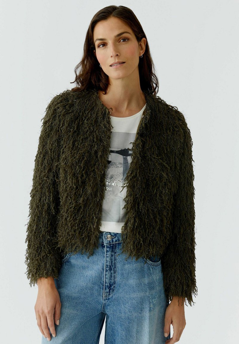 Olive green, shaggy-textured jacket with a cropped design, featuring a round neckline and open front, worn over a graphic t-shirt and jeans.