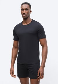 Άνδρας που στέκεται φορώντας εφαρμοστό μαύρο κοντομάνικο t-shirt και μαύρα σορτς μπροστά σε απλό φωτεινό φόντο.