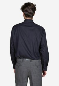 Donkerblauw overhemd met lange mouwen, gecombineerd met grijs geruite broeken. Het overhemd heeft een klassieke kraag en knoopmanchetten; de broeken hebben achterzakken en riemlussen.