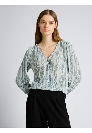 Jonge vrouw met steil bruin haar, gekleed in een lichtblauw blouse met patroon en opgeblazen mouwen, en zwarte hoge taille broek.