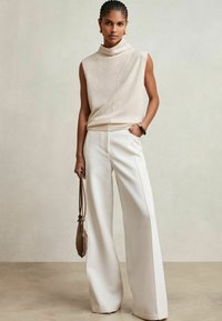 Beige ärmlös topp med hög halsringning, i kombination med vida vita byxor. Håller en brun väska. Slät textur, minimalistisk design.
