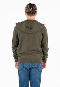 Felpa con cappuccio verde oliva, con orli e polsini a coste, caratterizzata da una texture sottile e una tasca posteriore. Indossata con jeans blu.