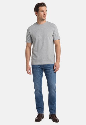 Mann steht, trägt ein hellgraues kurzärmeliges T-Shirt, blaue Jeans und dunkelbraune Schuhe, blickt nach rechts.