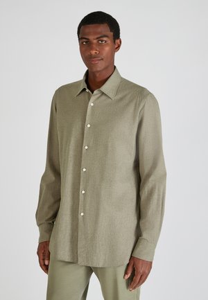 Man in een licht olijfgroene knopshirt met lange mouwen en bijpassende broek, staande tegen een effen lichtgrijze achtergrond.