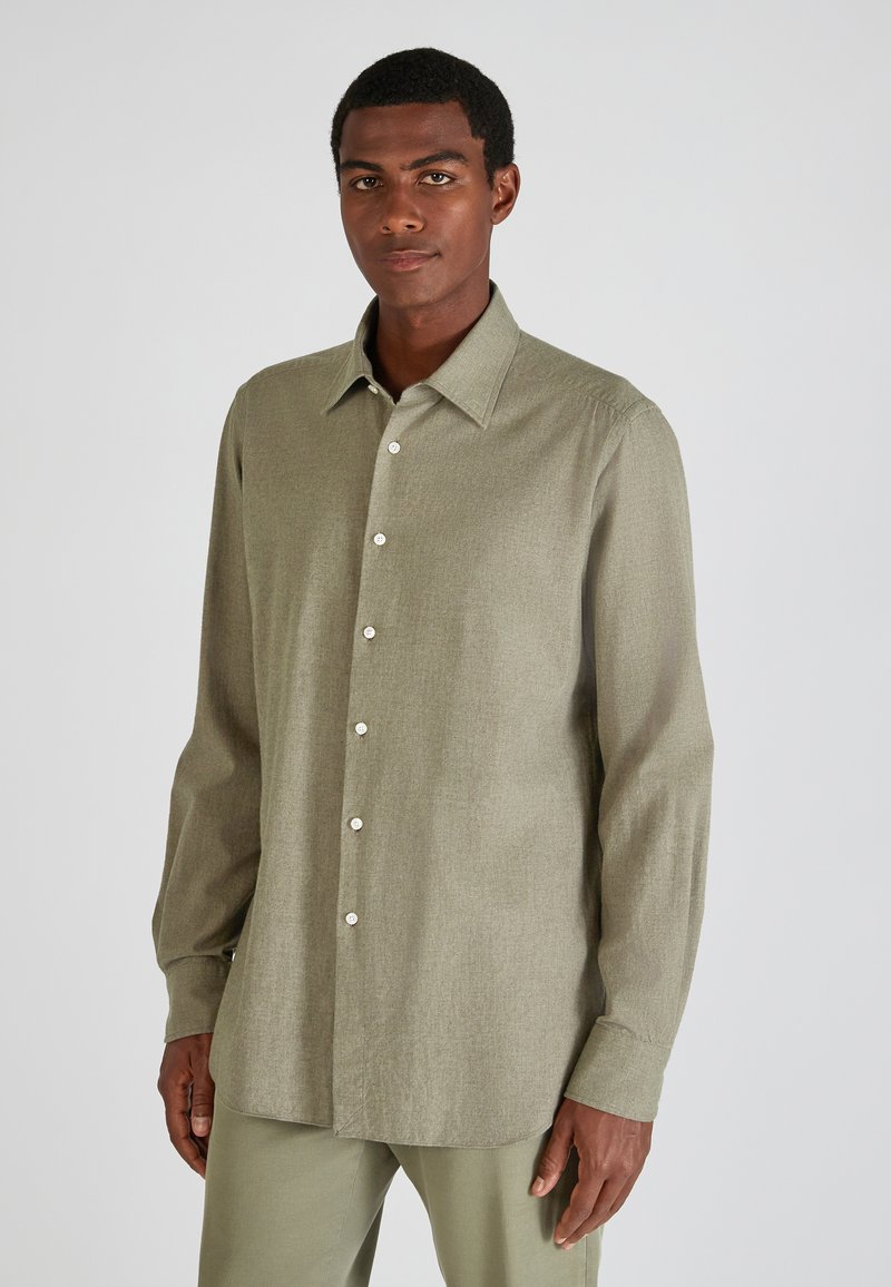 Man in een licht olijfgroene knopshirt met lange mouwen en bijpassende broek, staande tegen een effen lichtgrijze achtergrond.