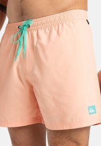 Shorts de bain pêche clair avec une ceinture élastique, un cordon de serrage teal, une poche latérale et un petit badge logo sur la jambe inférieure gauche.