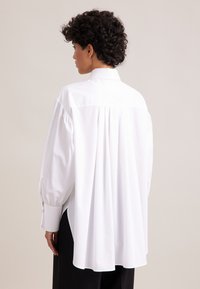 Weiße Bluse mit langen Ärmeln, plissiertem Rücken, spitzen Kragen und knöpfbaren Manschetten. Glattes Material mit lockerer Passform und seitlichen Schlitzen am Saum.