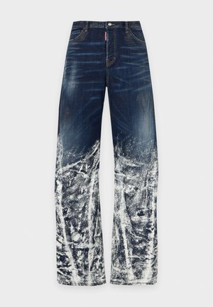 Modèle portant une veste en jean, une chemise blanche, un jean large effet usé et des sandales, avec un sac à main bleu en jean sur un fond uni.