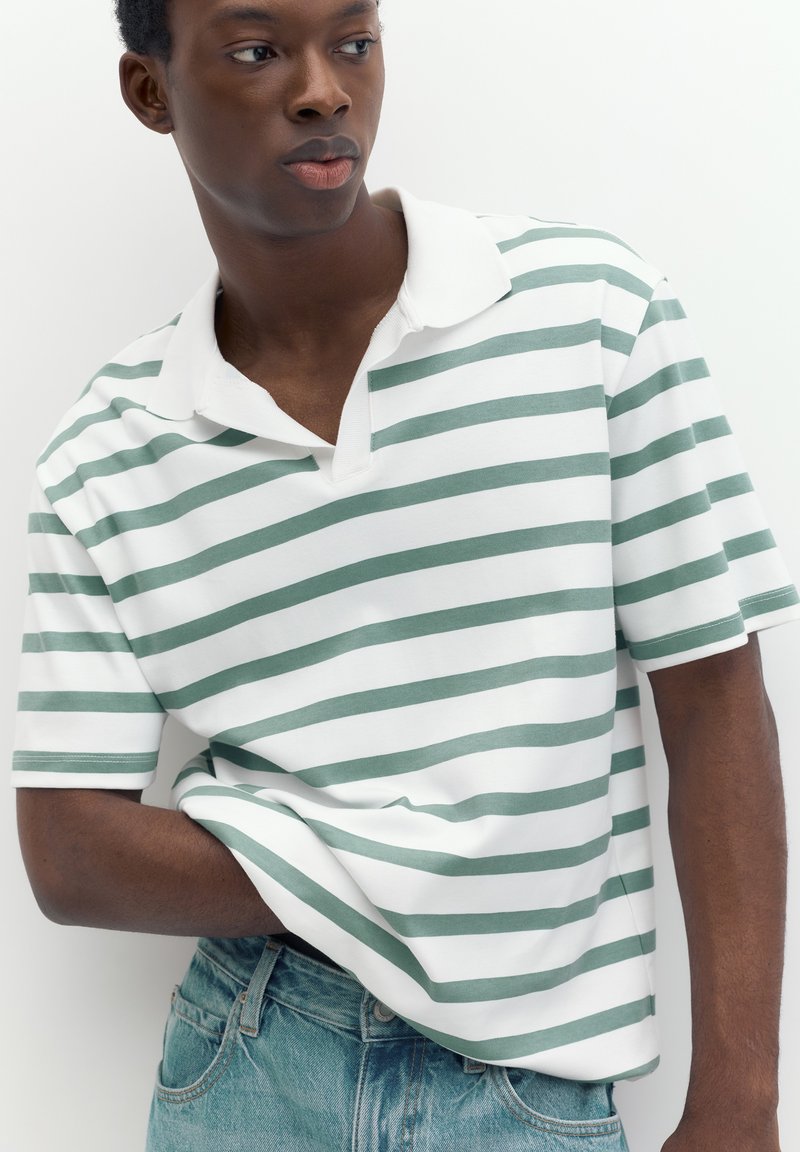 Jeune homme portant un polo blanc et vert rayé, une main dans la poche de son jean, regardant sur le côté sur fond blanc.